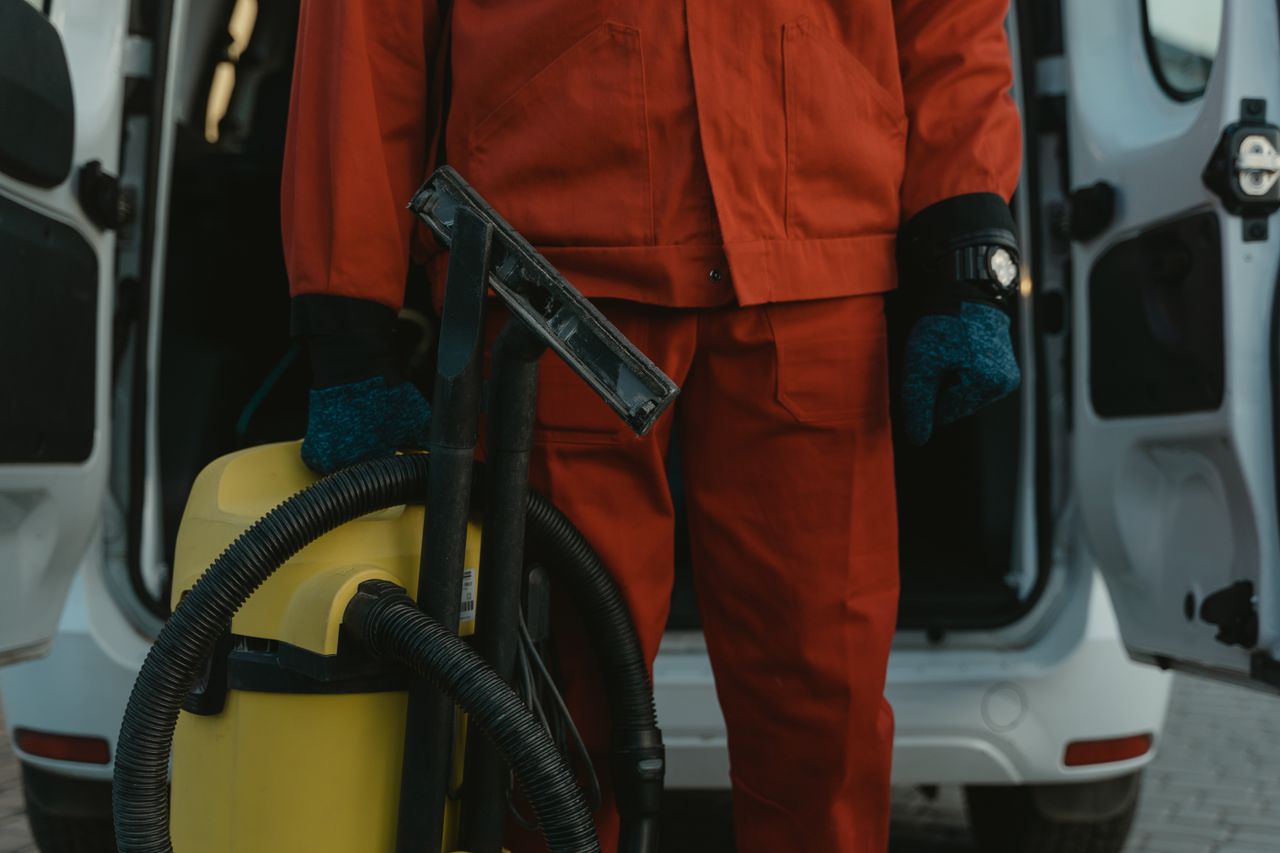 A skilled cleaner wearing a red uniform stands beside a van with a vacuum, prepared to begin service.